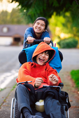 Big sister pushing disabled little brother in wheelchair around neighborhood, laughing and smiling