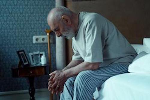 An old man with Dimensia, seated on his bed, staring into space